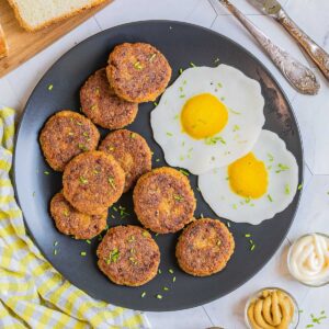 Nine round flat crispy brown sausage patties on a black plate served with fried egg look-alikes. Condiments, bread, utensils, and a glass of juice is placed next to it.