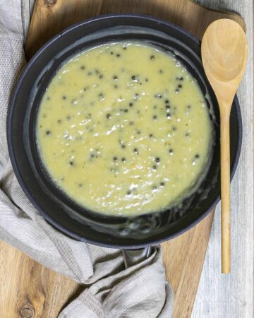 Black frying pan with a light green sauce with whole peppercorns. A wooden spoon is placed next to it.