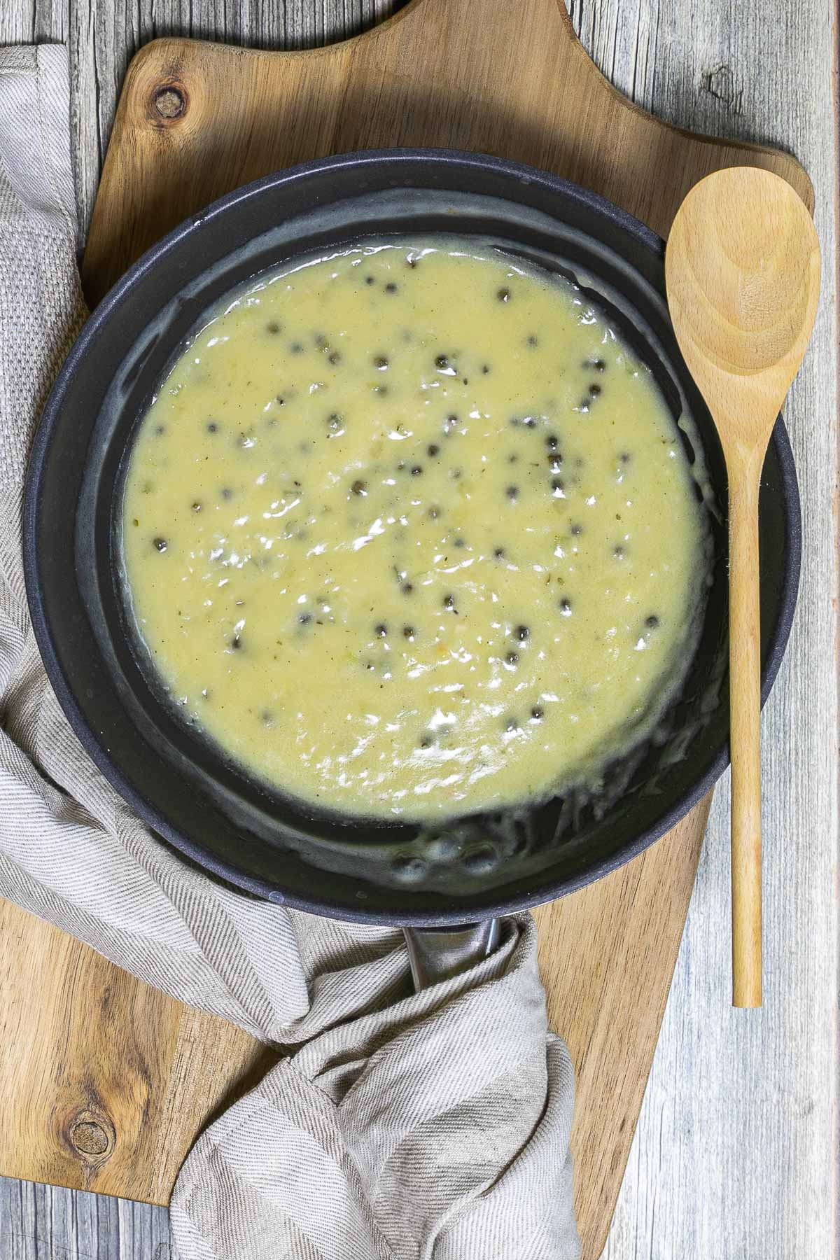 Black frying pan with a light green sauce with whole peppercorns. A wooden spoon is placed next to it. 