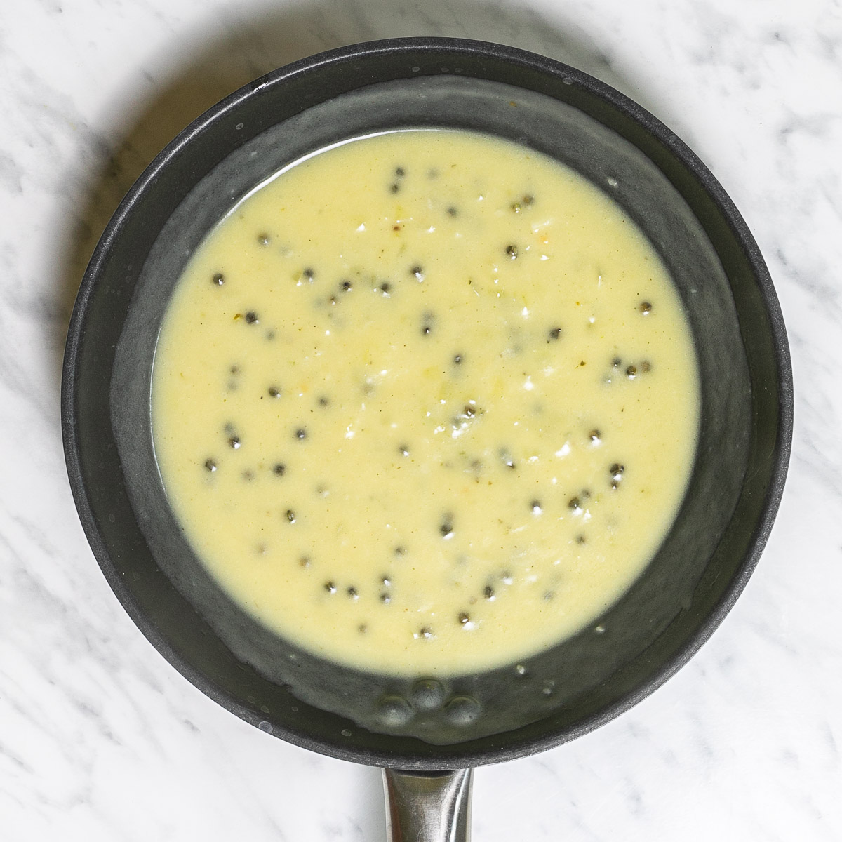 Black frying pan with a light green sauce with whole peppercorns.