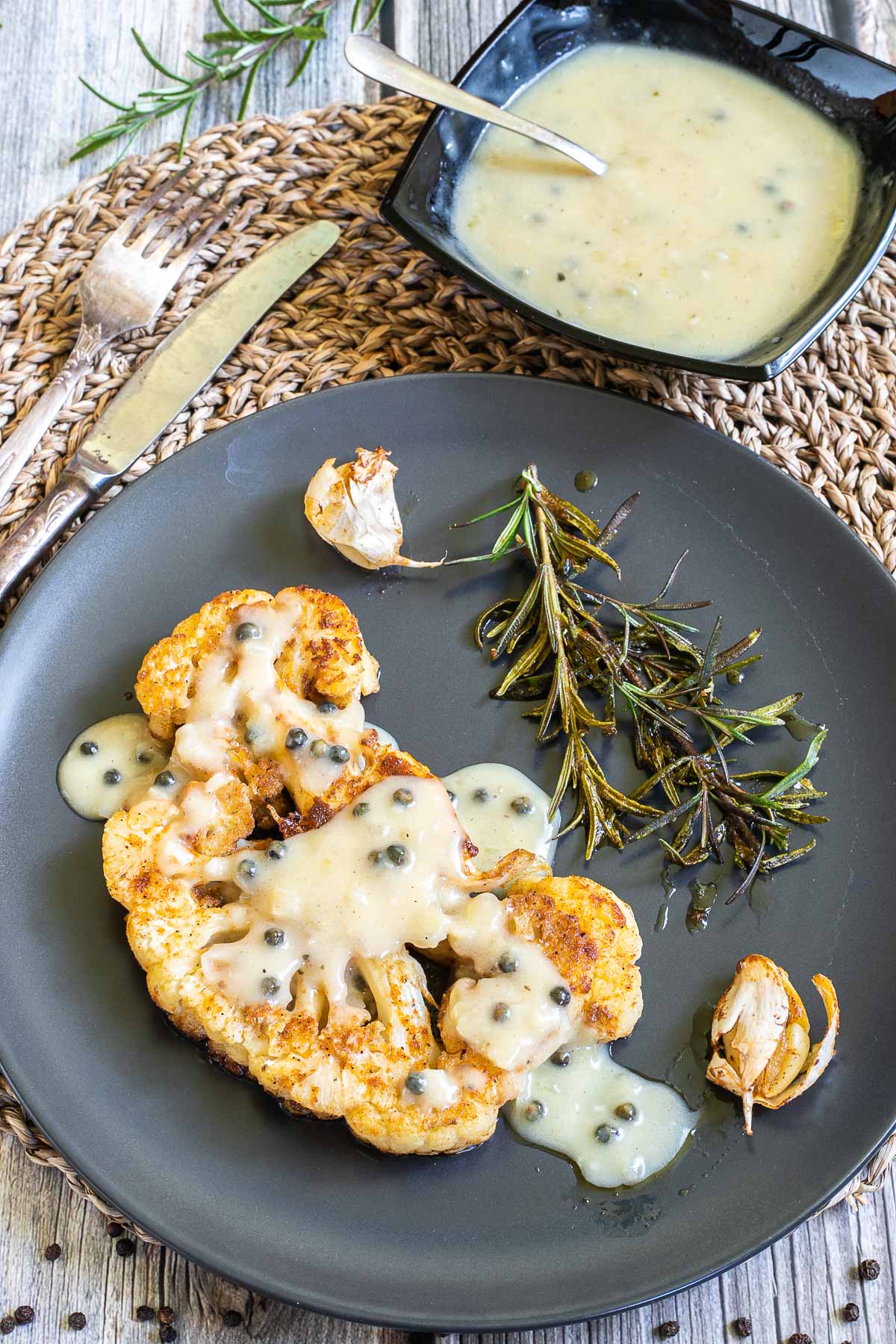 Black plate with a large slice of cauliflower drizzled with green peppercorn sauce. Roasted garlic and rosemary are placed next to it. There is a small black bowl of the remaining sauce. 