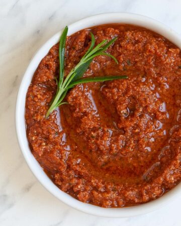 Red pesto with rosemary leaves in a small white bowl.