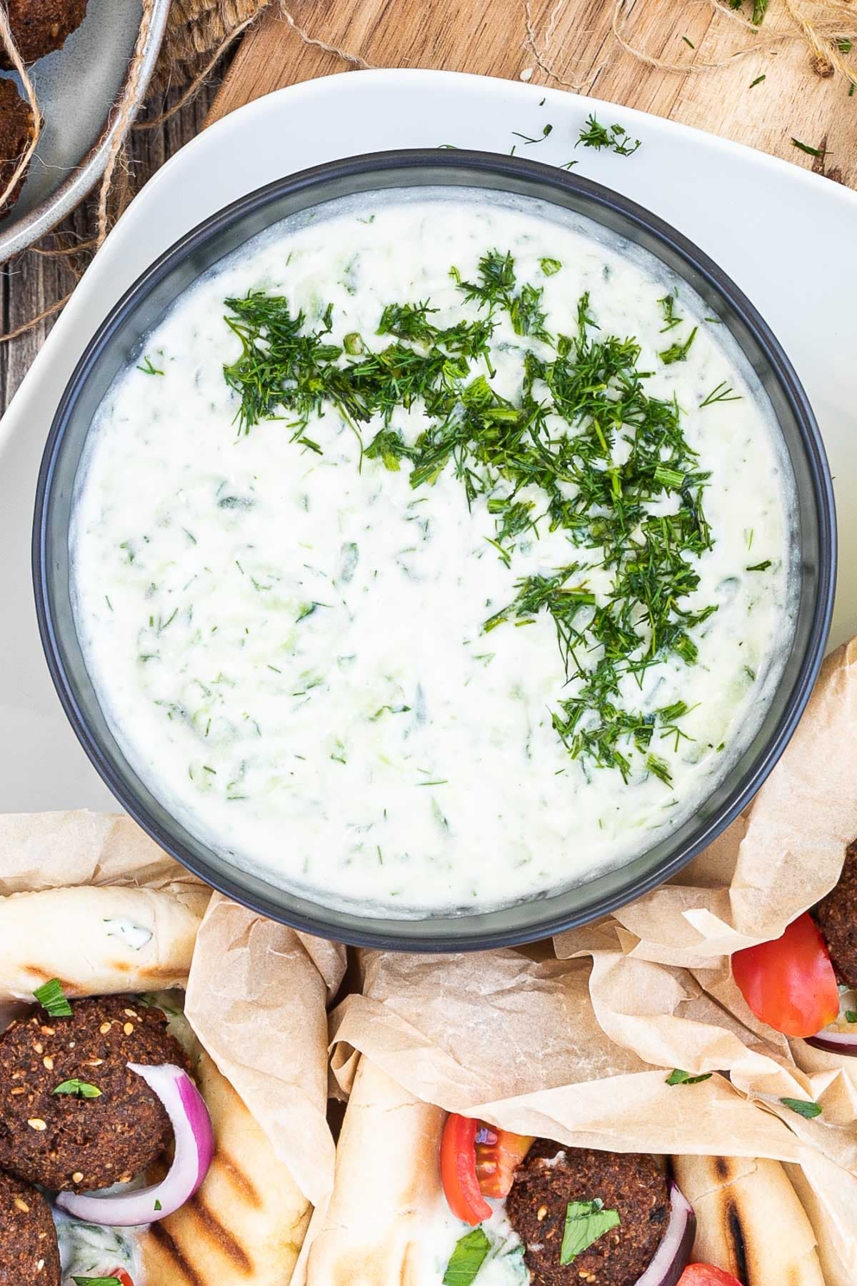 Black bowl with white green sauce topped with freshly chopped dill. The bowl is surrounded by falafel in pita bread on a white plate.