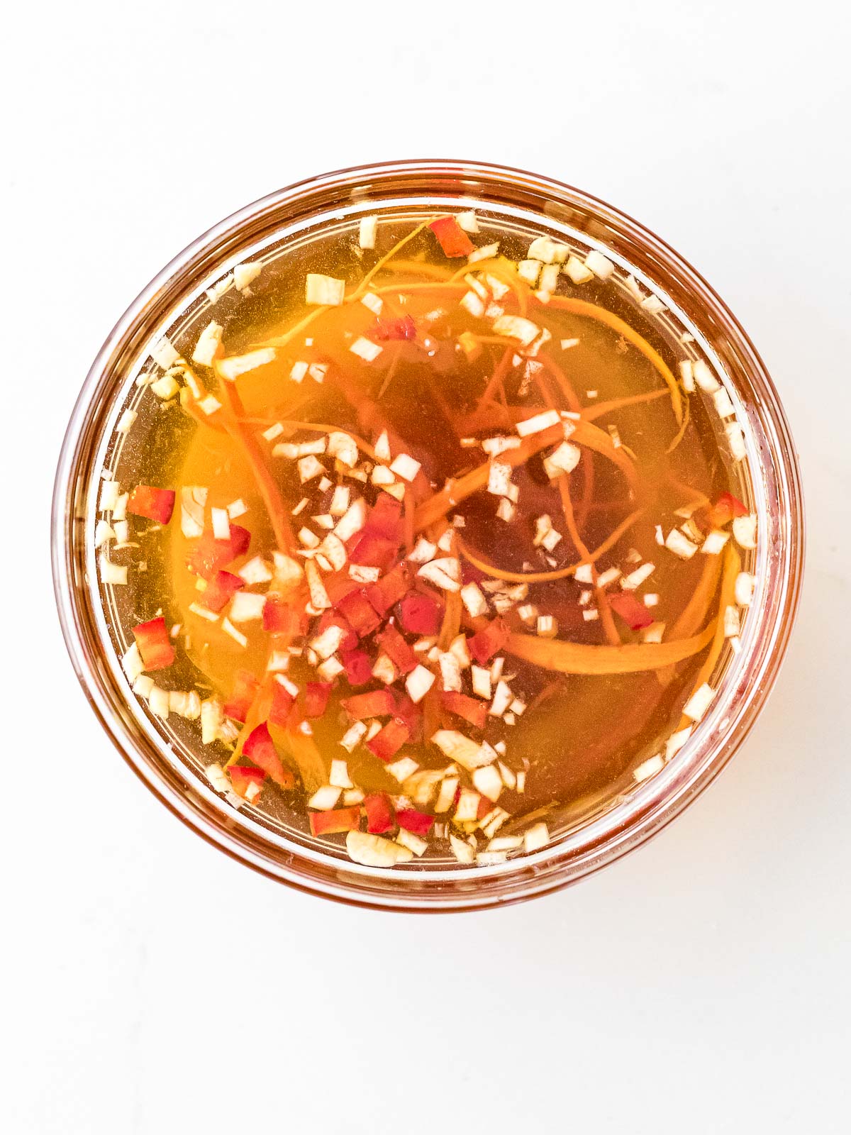 A light golden brown thin sauce in a small galss bowl with chunks of ginger, chili pepper and carrot shreds swimming on the surface