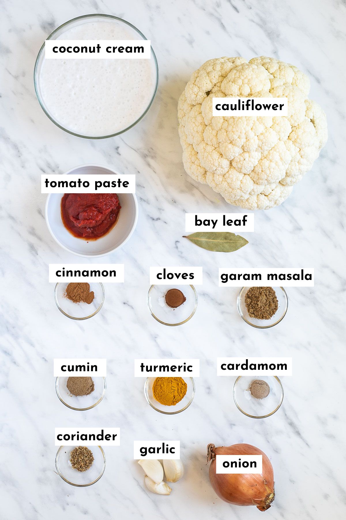 Ingredients of cauliflower korma in glass bowls like coconut cream, tomato paste, various spices and a head of cauliflower next to them.