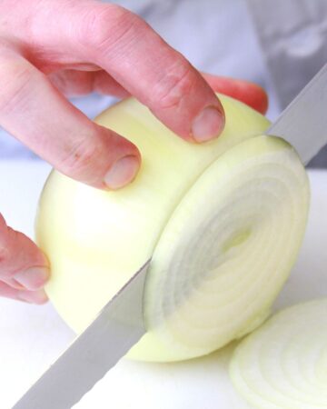 A hand is holding a large peeled onion and cutting it into slices with a large knife.