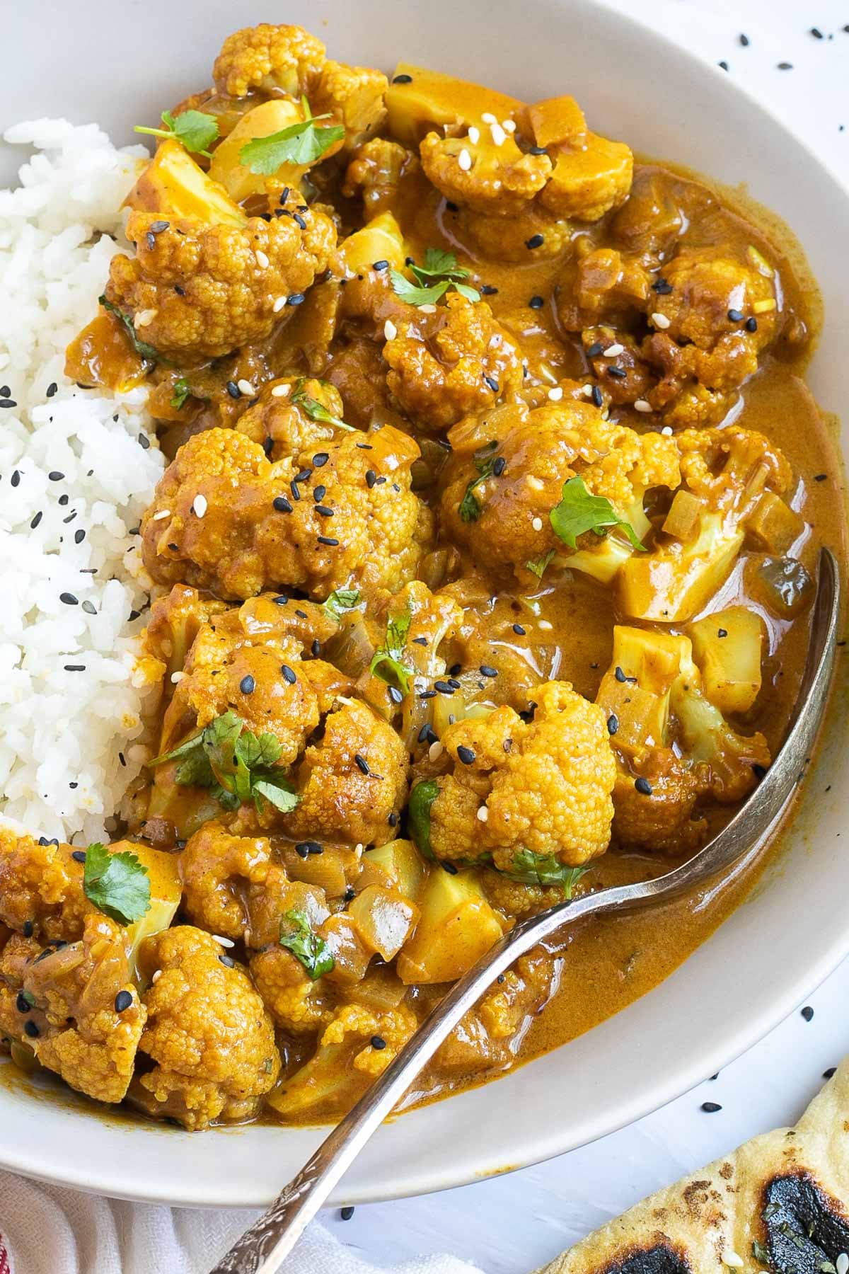 A white plate with white rice and cauliflower florets in a creamy orange sauce sprinkled freshly chopped cilantro. A spoon is placed inside.