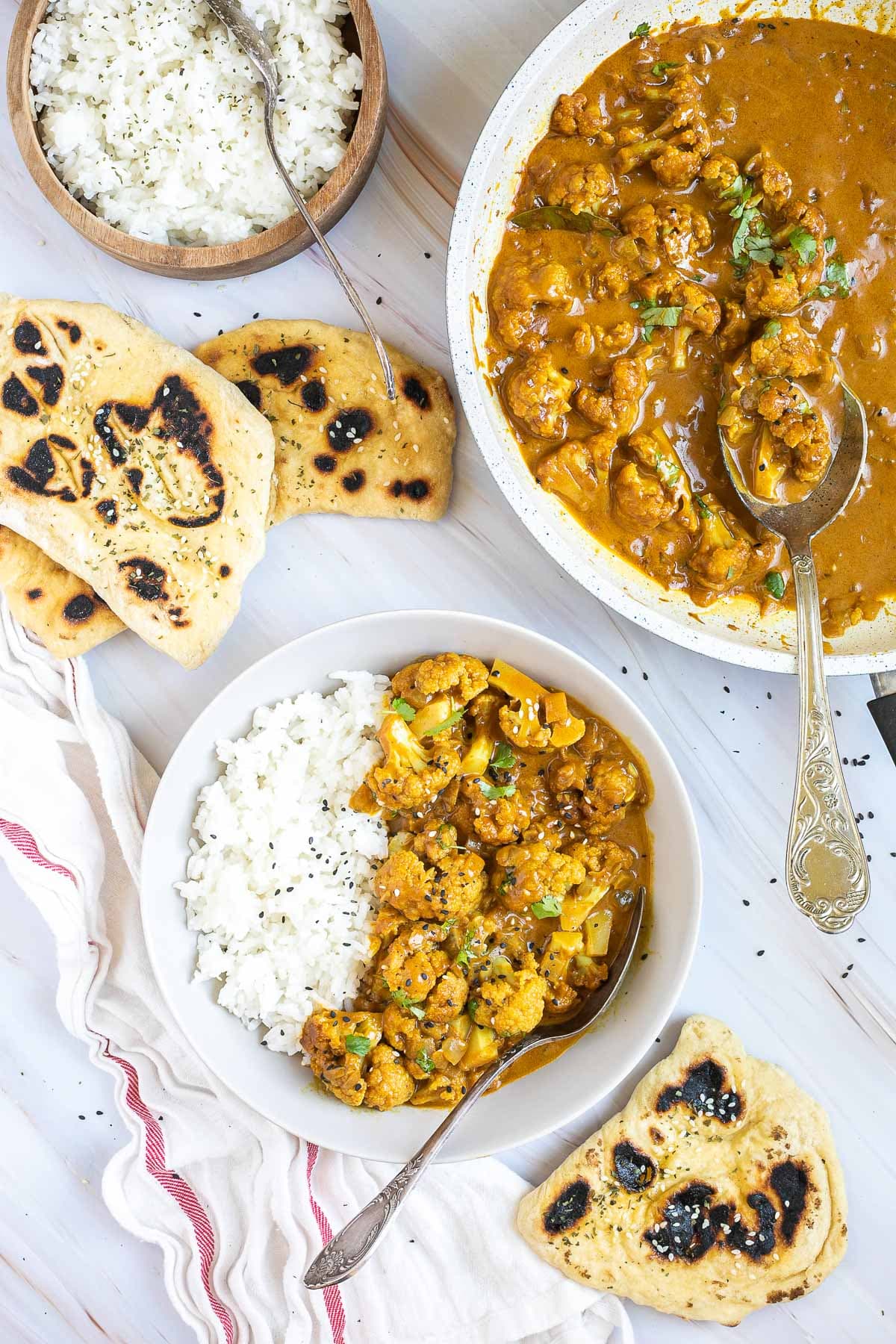 A white plate with white rice and cauliflower florets in a creamy orange sauce sprinkled freshly chopped cilantro. A spoon is placed inside. Naan bread, a bowl of rice, and leftover cauliflower korma are next to it.
