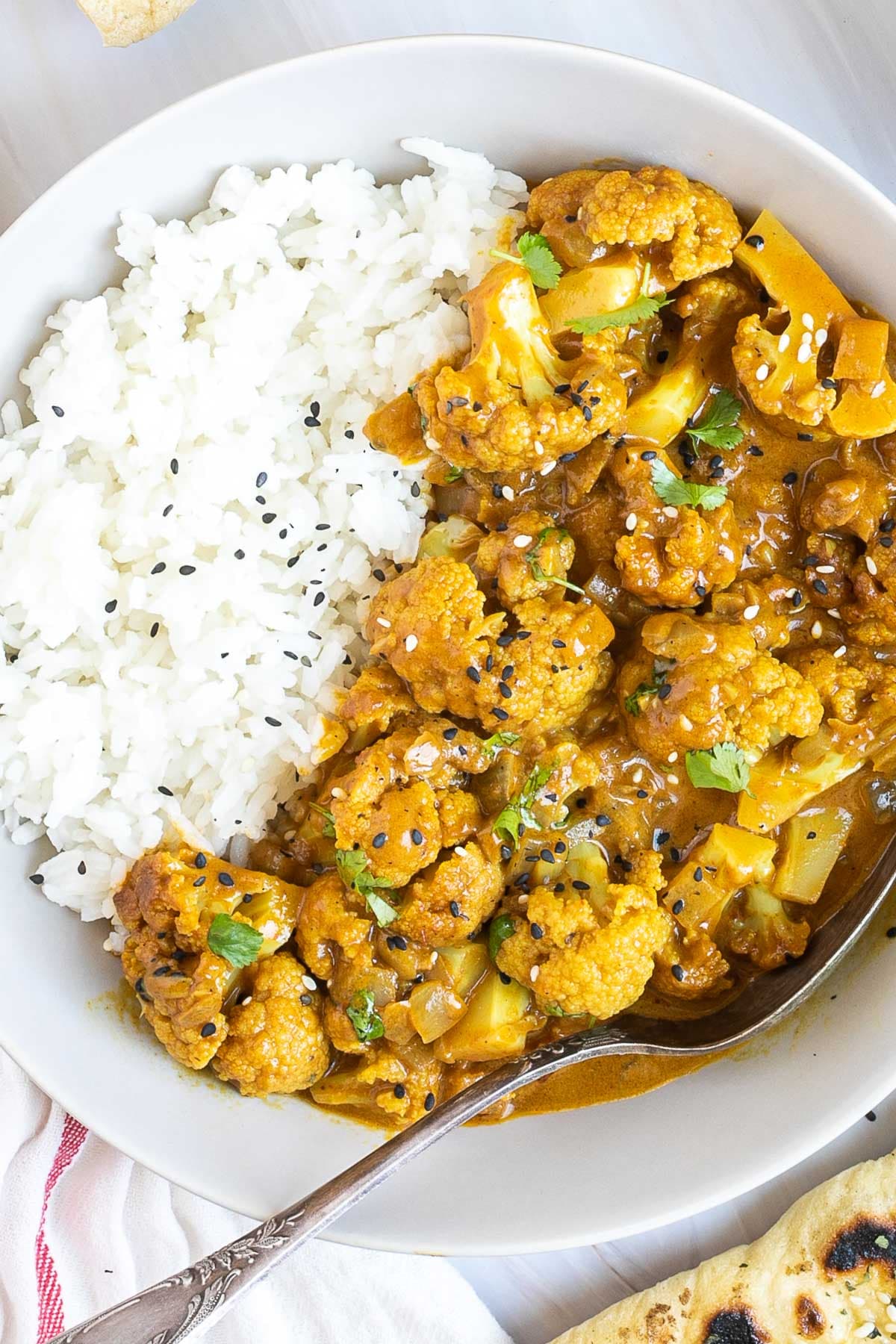 A white plate with white rice and cauliflower florets in a creamy orange sauce sprinkled freshly chopped cilantro. A spoon is placed inside.