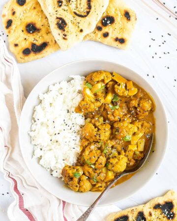 A white plate with white rice and cauliflower florets in a creamy orange sauce sprinkled freshly chopped cilantro. A spoon is placed inside. Naan bread pieces are next to it.