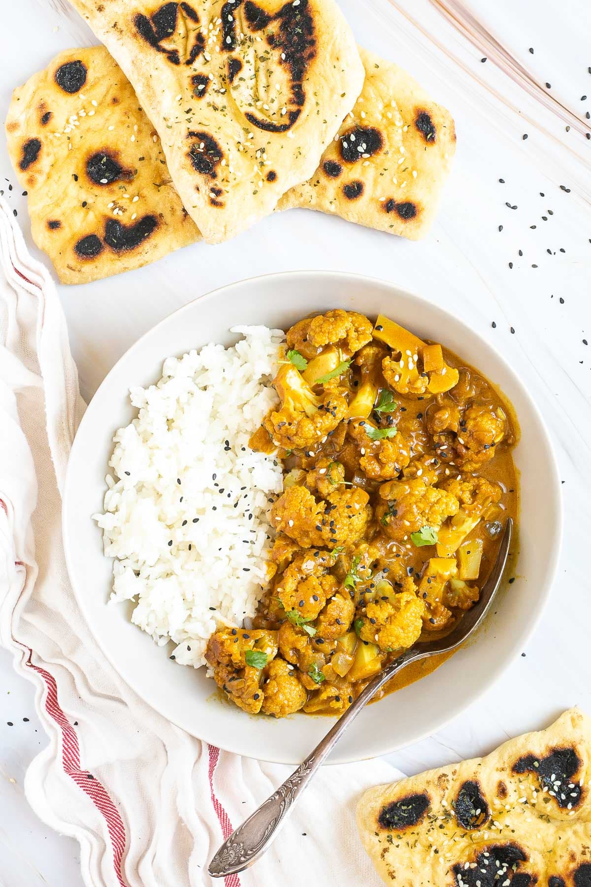 A white plate with white rice and cauliflower florets in a creamy orange sauce sprinkled freshly chopped cilantro. A spoon is placed inside. Naan bread pieces are next to it.