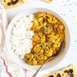 A white plate with white rice and cauliflower florets in a creamy orange sauce sprinkled freshly chopped cilantro. A spoon is placed inside. Naan bread pieces are next to it.