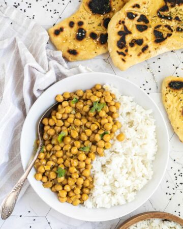 A white plate with white rice and chickpeas in a creamy orange sauce sprinkled freshly chopped cilantro. A spoon is placed inside. Naan bread and a bowl of rice are next to it.
