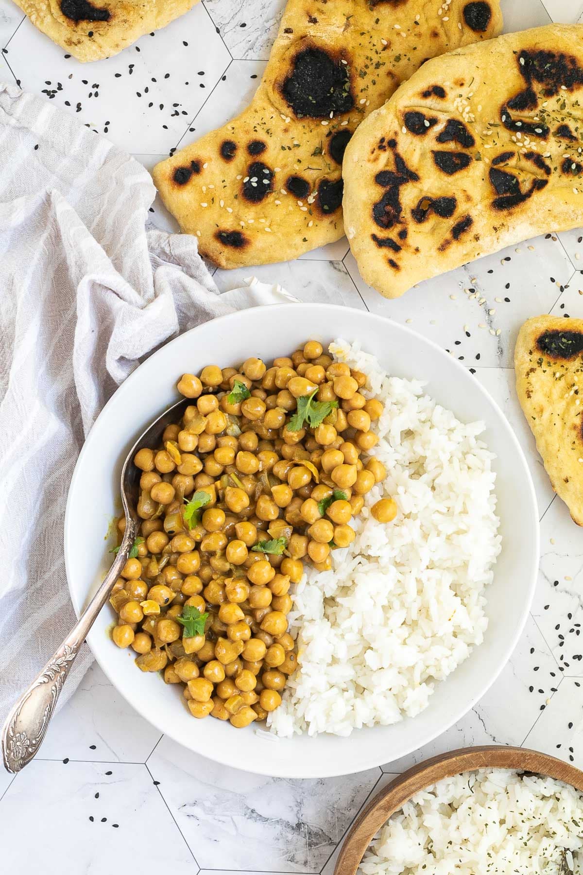A white plate with white rice and chickpeas in a creamy orange sauce sprinkled freshly chopped cilantro. A spoon is placed inside. Naan bread and a bowl of rice are next to it.