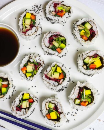 Large white round plate with 11 rainbow sushi rolls without fish or seafood, only vegetables. Served with a small bowl of soy sauce. Blue chopsticks are placed next to it.