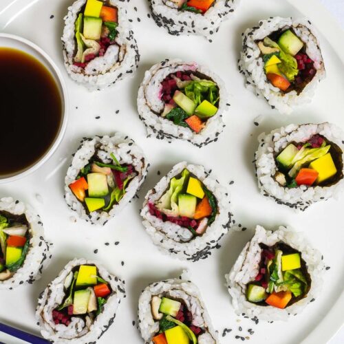Large white round plate with 11 rainbow sushi rolls without fish or seafood, only vegetables. Served with a small bowl of soy sauce. Blue chopsticks are placed next to it.