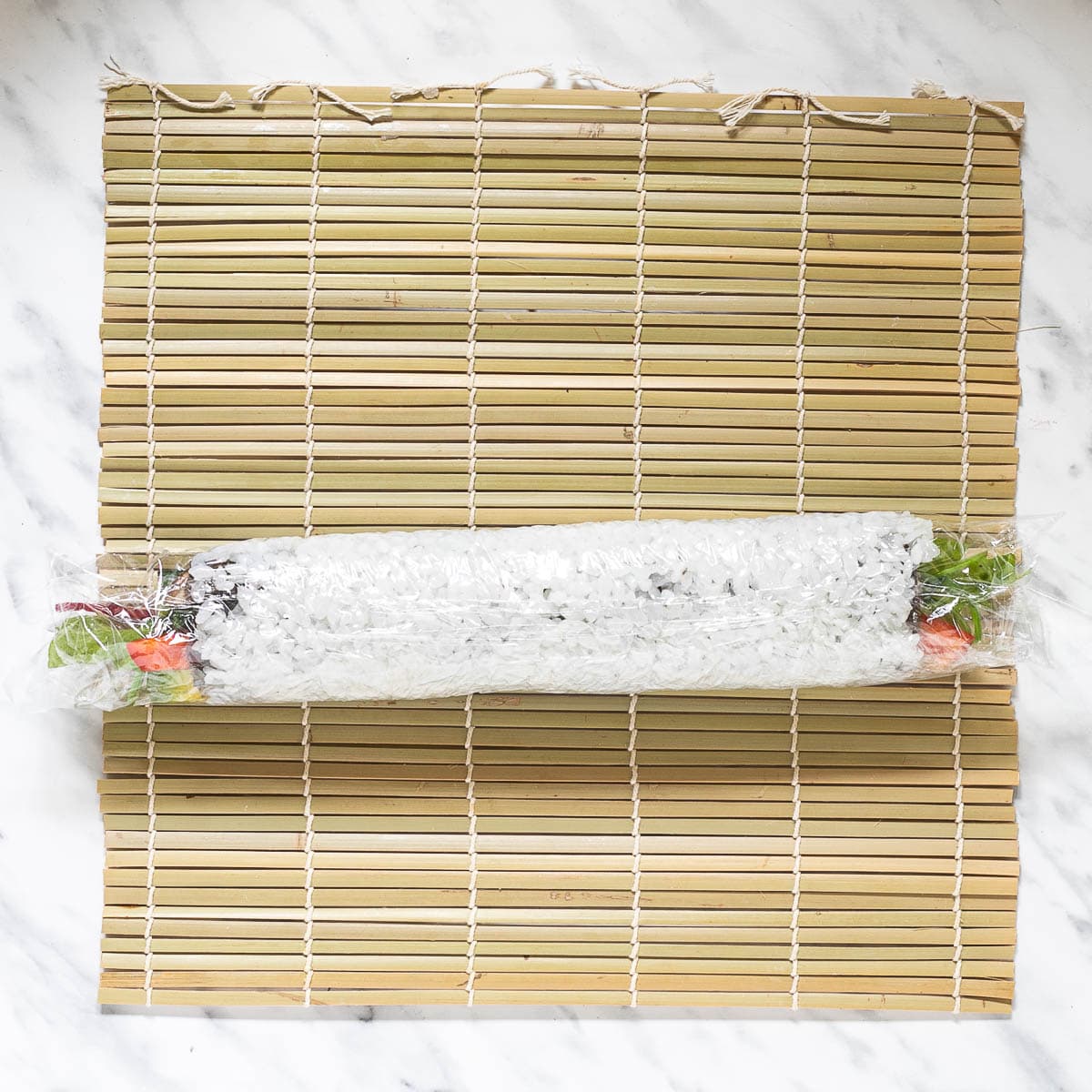 A long sushi roll wrapped in plastic wrap on top of a bamboo mat.