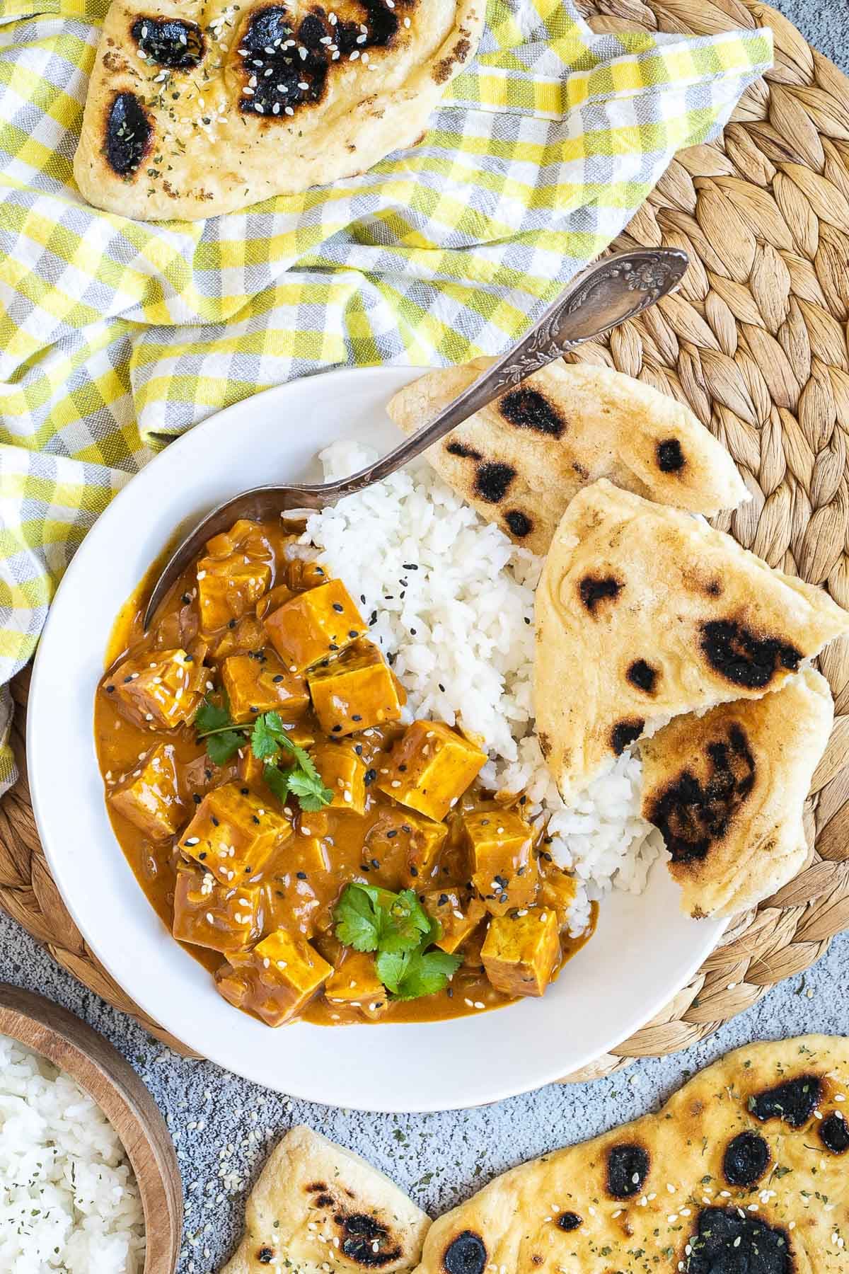 A white plate with white rice, pieces of naan bread and tofu cubes in a creamy orange sauce sprinkled with sesame seeds and freshly chopped cilantro. A spoon is placed inside. 