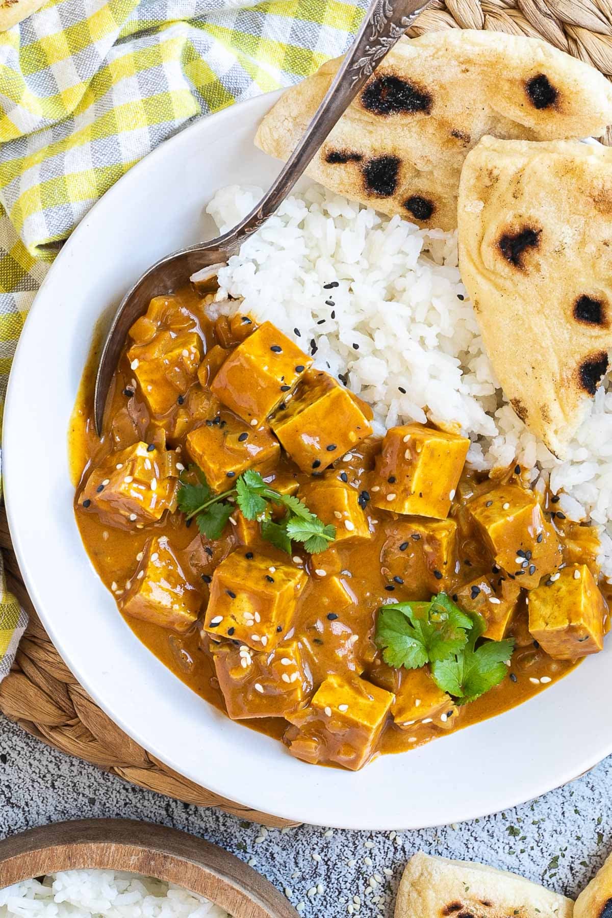 A white plate with white rice, pieces of naan bread and tofu cubes in a creamy orange sauce sprinkled with sesame seeds and freshly chopped cilantro. A spoon is placed inside.