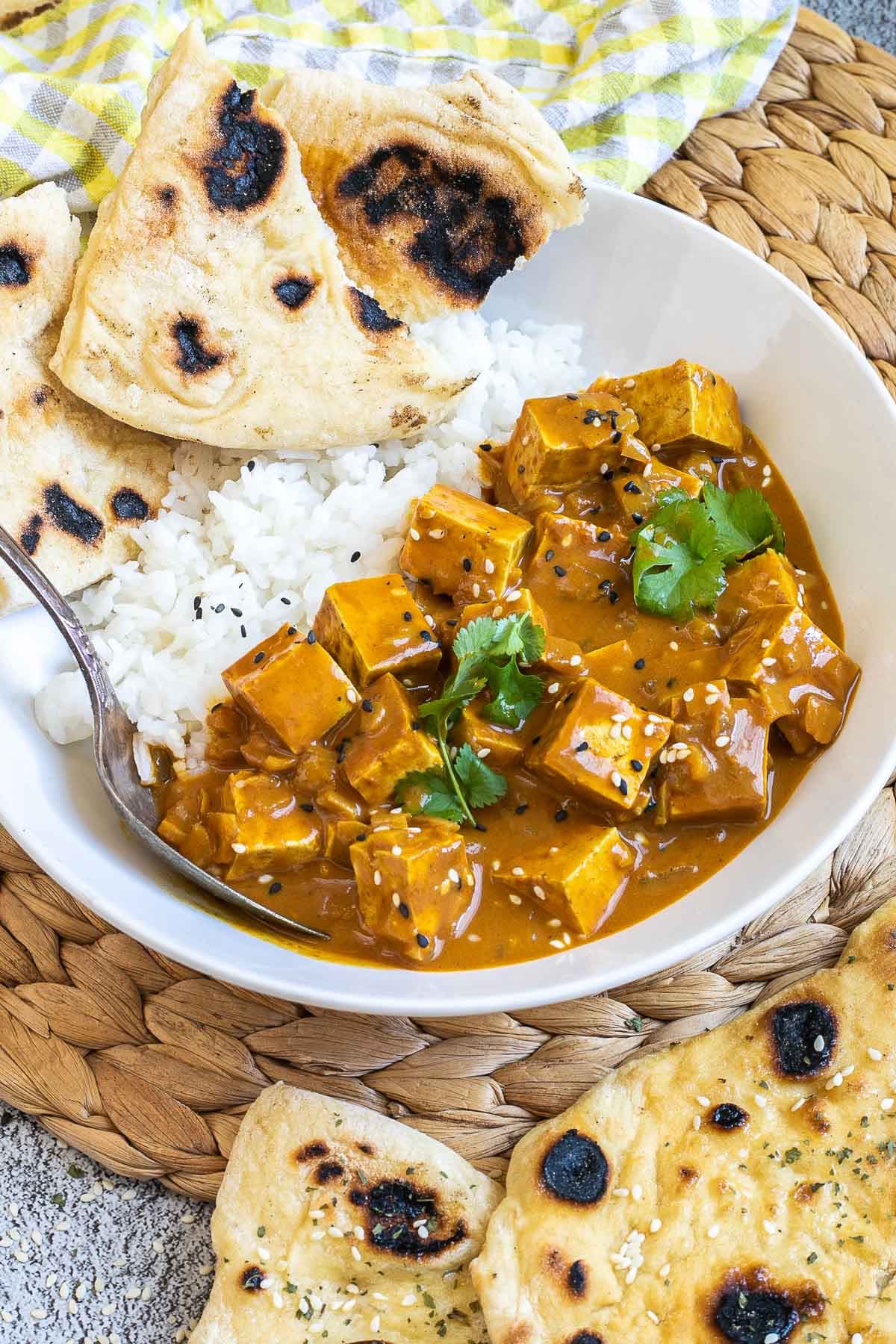 A white plate with white rice, pieces of naan bread and tofu cubes in a creamy orange sauce sprinkled with sesame seeds and freshly chopped cilantro. A spoon is placed inside.