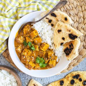 A white plate with white rice, pieces of naan bread and tofu cubes in a creamy orange sauce sprinkled with sesame seeds and freshly chopped cilantro. A spoon is placed inside.