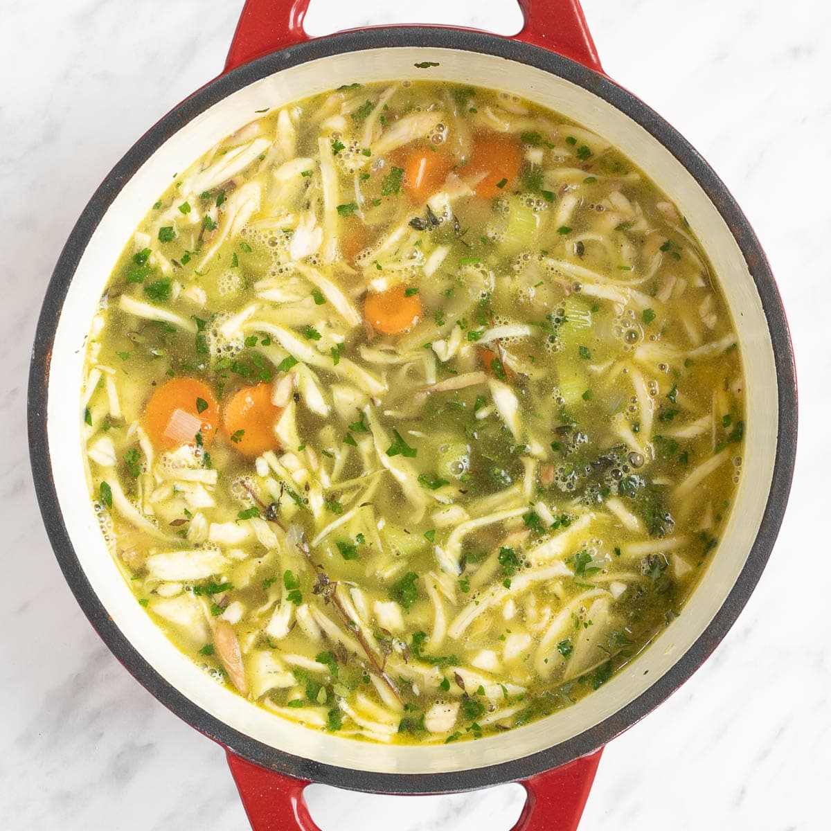 A red-white enameled Dutch oven with chopped veggies like carrot, celery, and onion, shredded mushroom pieces, green herbs in vegetable broth.