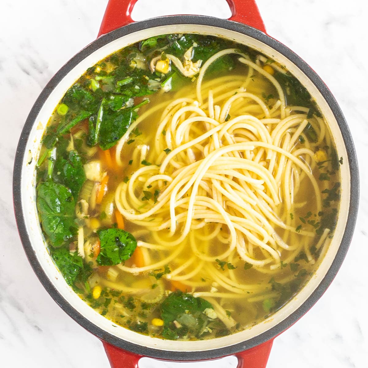 Red Dutch oven with sliced carrots, chopped parsnip, chopped celeriac, celery stalk, shredded oyster mushrooms, chopped herbs, fresh spinach leaves, corn, green peas, and noodles in a thin vegetable broth-like soup.