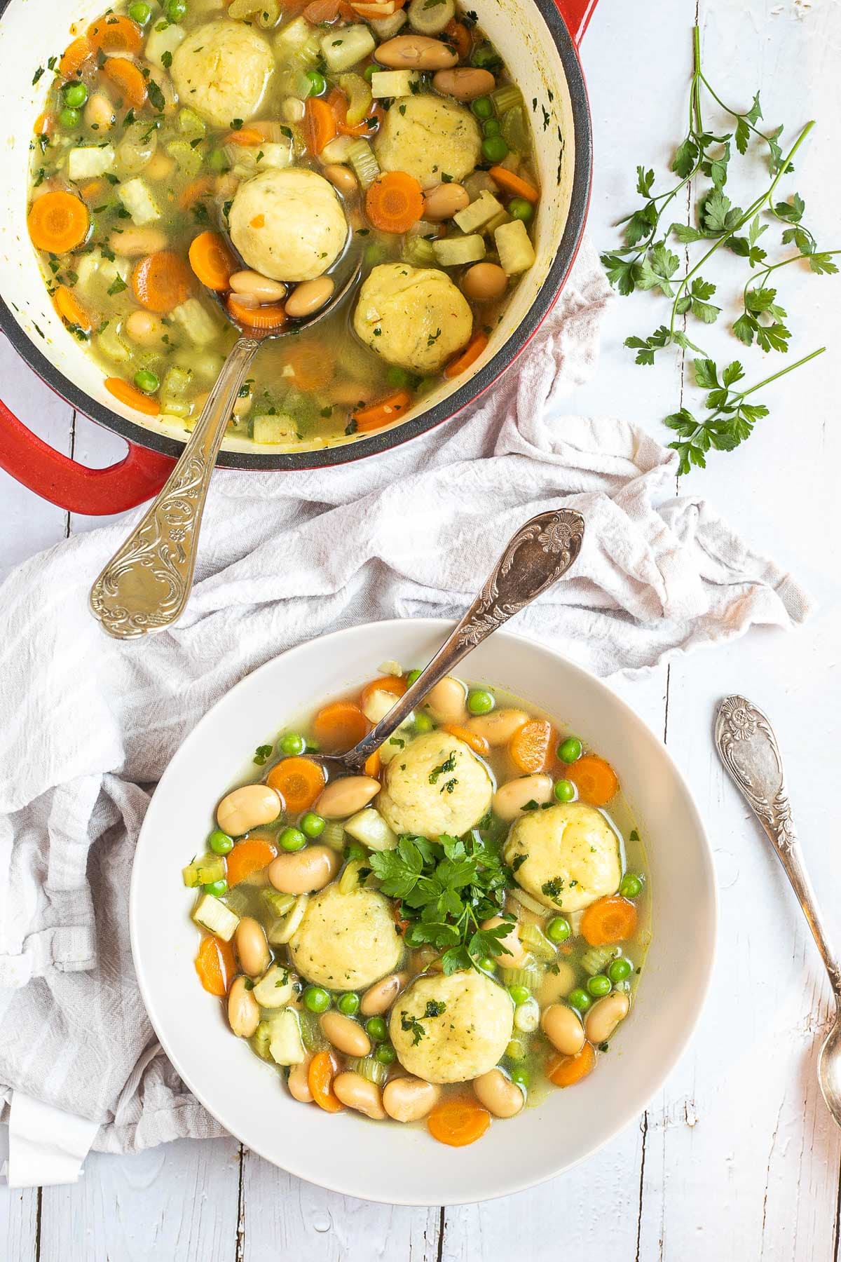 A white plate with dumplings, chopped veggies, green herbs, white beans, and green peas in a vegetable broth soup. A spoon is placed in it. The remaining soup is in a red white enameled Dutch oven next to it. 