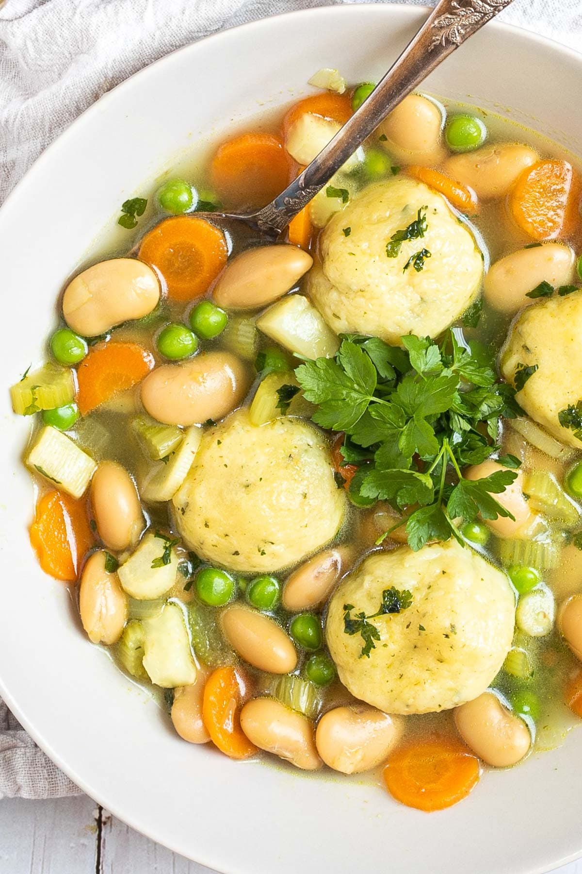 A white plate with dumplings, chopped veggies, green herbs, white beans, and green peas in a vegetable broth soup. A spoon is placed in it.