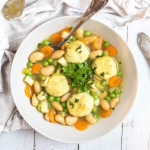 A white plate with dumplings, chopped veggies, green herbs, white beans, and green peas in a vegetable broth soup. A spoon is placed in it.