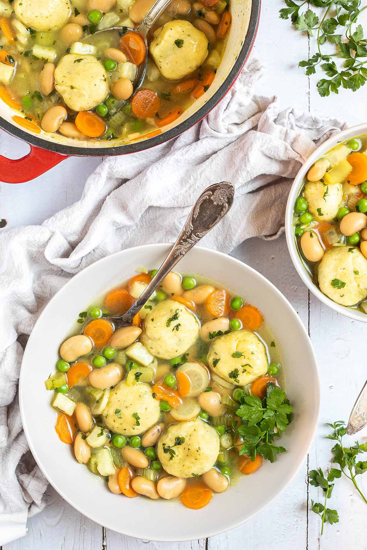 Two white plates with dumplings, chopped veggies, green herbs, white beans, and green peas in a vegetable broth soup. A spoon is placed in it. The remaining soup is in a red white enameled Dutch oven next to it.