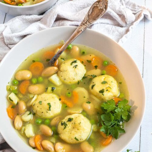 Two white plates with dumplings, chopped veggies, green herbs, white beans, and green peas in a vegetable broth soup. A spoon is placed in it.