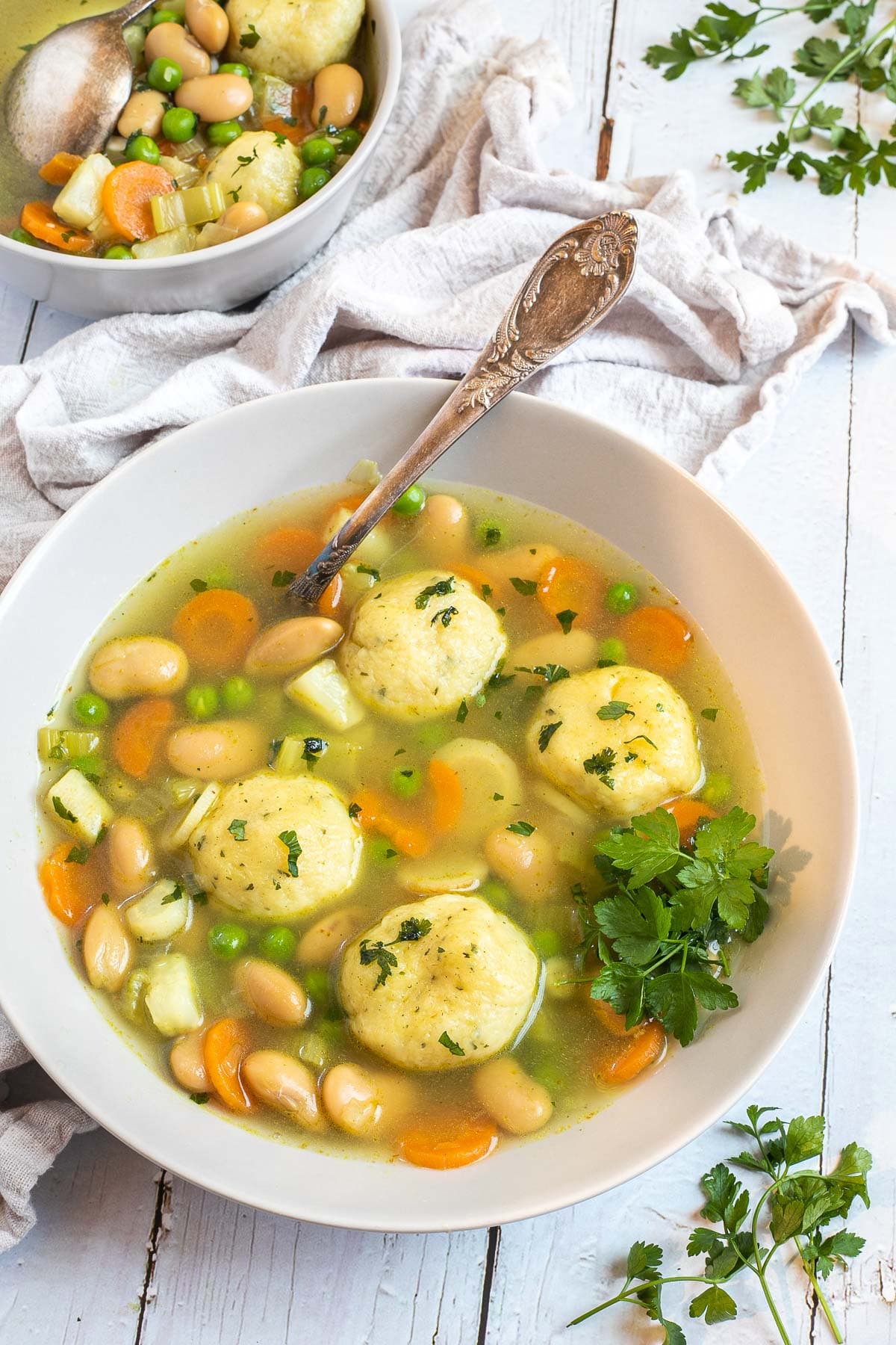 Two white plates with dumplings, chopped veggies, green herbs, white beans, and green peas in a vegetable broth soup. A spoon is placed in it.