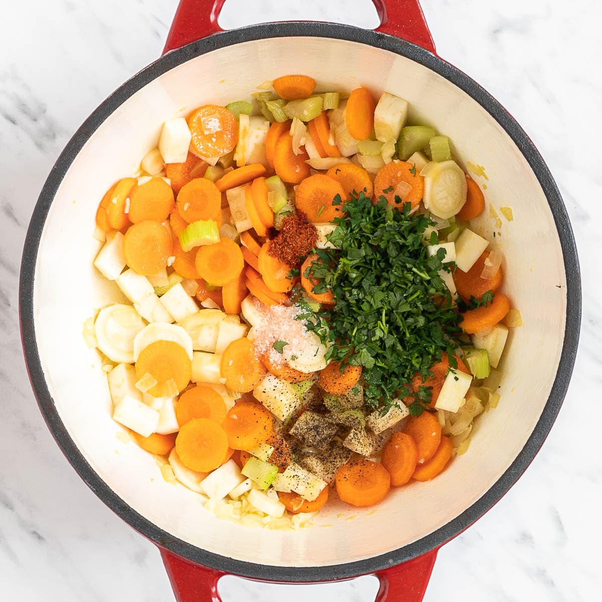 Red white enameled Dutch oven with chopped carrots, celery stalks, celeriac root, parsnip, and a large bunch of fresh green herbs,