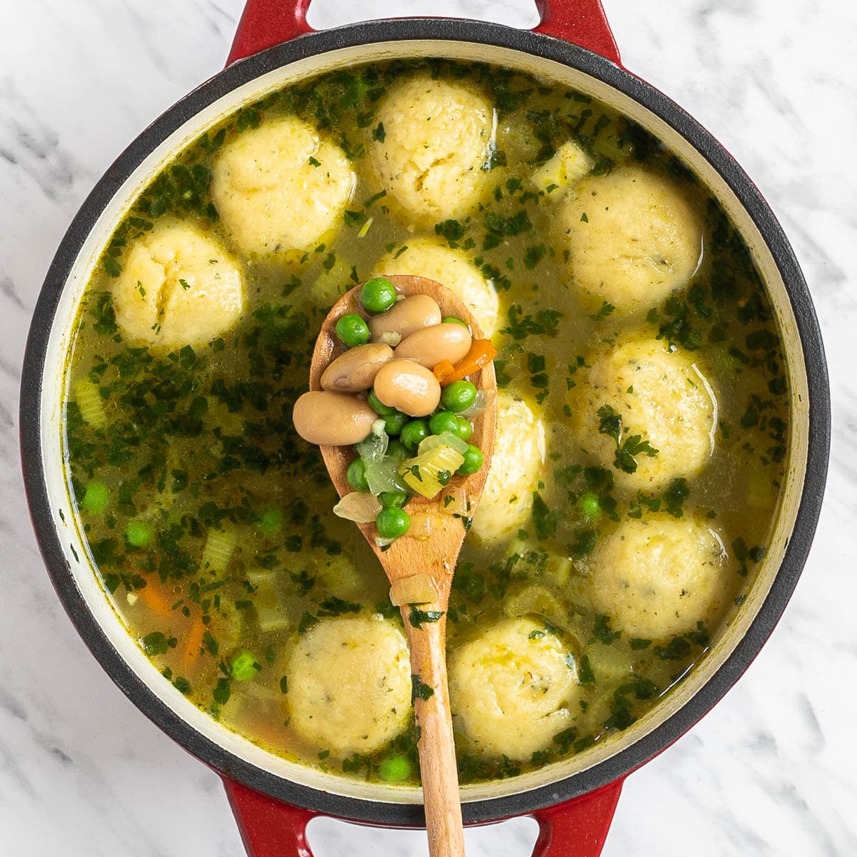 Dutch oven with dumplings, chopped veggies and green herbs in a light vegetable broth. A wooden spoon is holding white beans and green peas to be added.