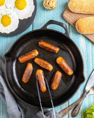 7 small sausages in a black cast-iron skillet. A hand holding tongs is taking one. Fake fried eggs, hoagie rolls, ketchup, and mustard are around it.