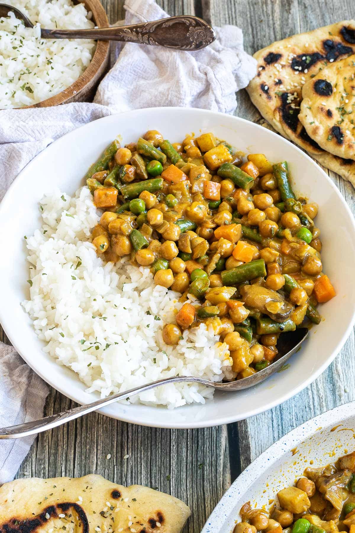 White plate with rice and different chopped vegetables, green peas, green beans, and chickpeas. Leftovers are next to it in the frying pan. Naan bread and more rice are on the side.