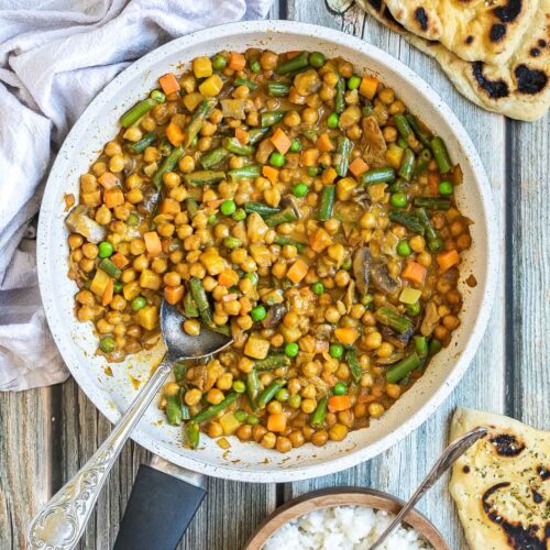 White frying pan full of vegetables like chickpeas, green beans, chopped carrots, green peas, chopped mushroom, and potatoes swimming in a brown-orange sauce. Naan bread and a brown bowl of rice are next to it.