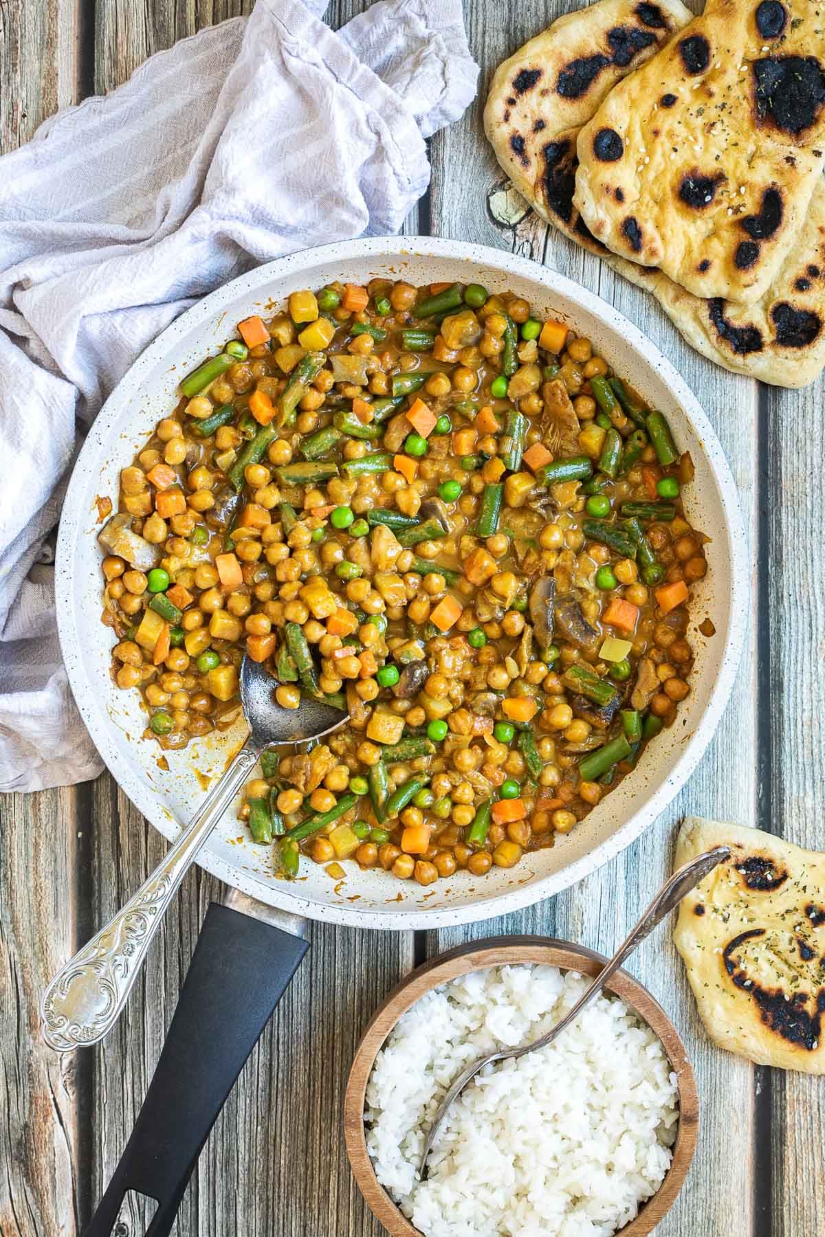 White frying pan full of vegetables like chickpeas, green beans, chopped carrots, green peas, chopped mushroom, and potatoes swimming in a brown-orange sauce. Naan bread and a brown bowl of rice are next to it.