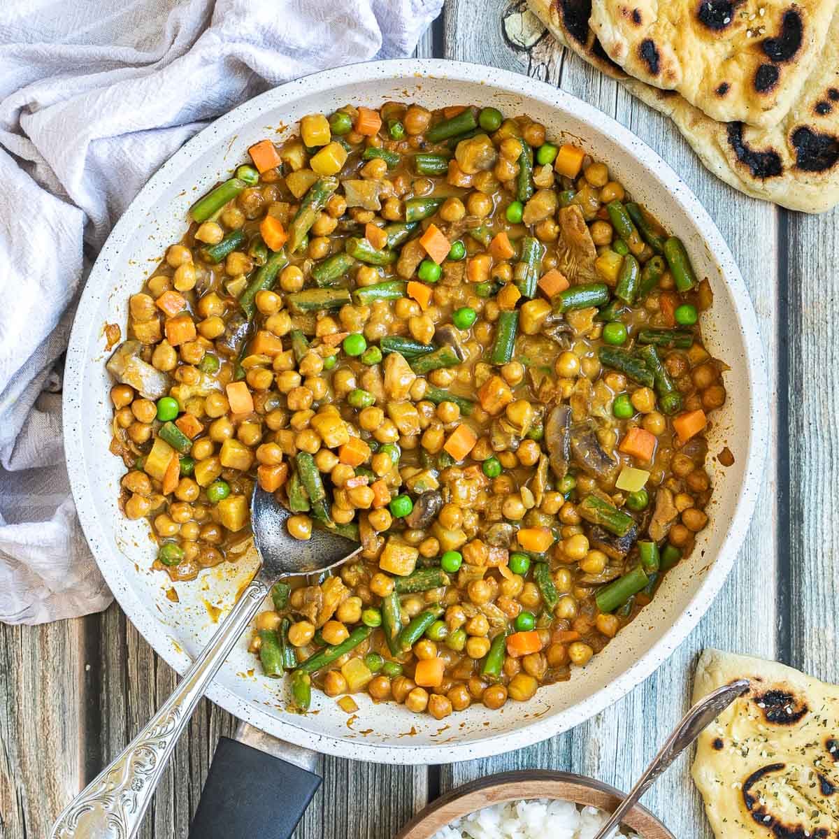 White frying pan full of vegetables like chickpeas, green beans, chopped carrots, green peas, chopped mushroom, and potatoes swimming in a brown-orange sauce. Naan bread and a brown bowl of rice are next to it.