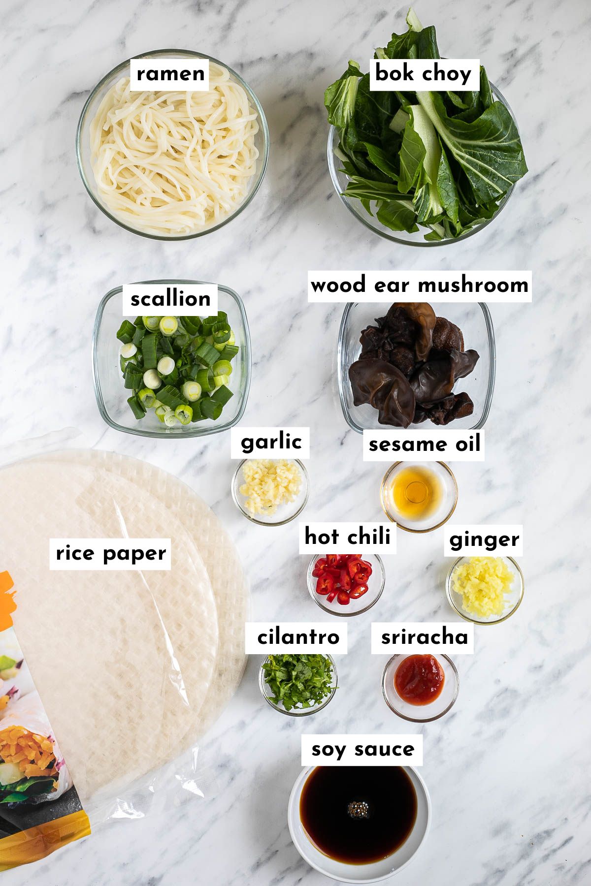 The ingredients of ramen spring rolls are placed in small glass bowls like ramen noodles, chopped bok choy, wood ear mushrooms, chopped scallion and cilantro, chopped garlic, red chili slices, minced ginger, hot sauce and oil. Dry rice paper wrappers are placed next to them.