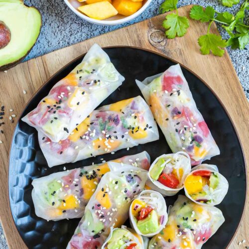 Fresh rice paper wraps filled with chopped tomato, mango, and avocado placed on a black plate. 4 half rolls and 6 whole rolls. Leftover ingredients are next to it.