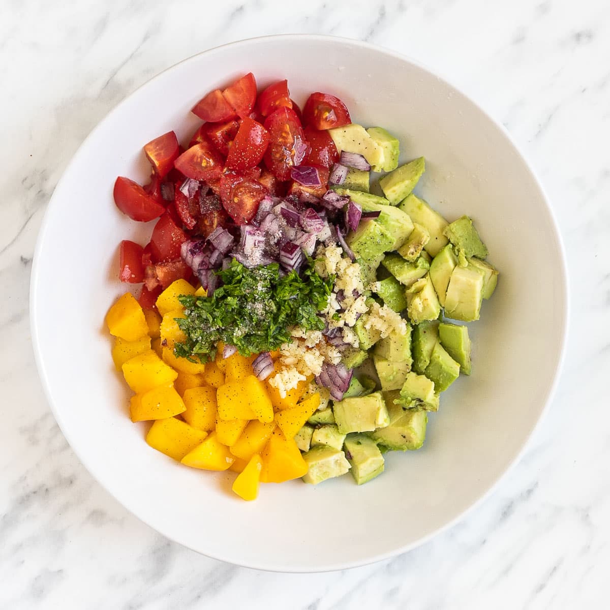 A white bowl of chopped green avocado, yellow mango, red cherry tomatoes, finely chopped red onion and chopped green herbs. Placed by color not mixed.