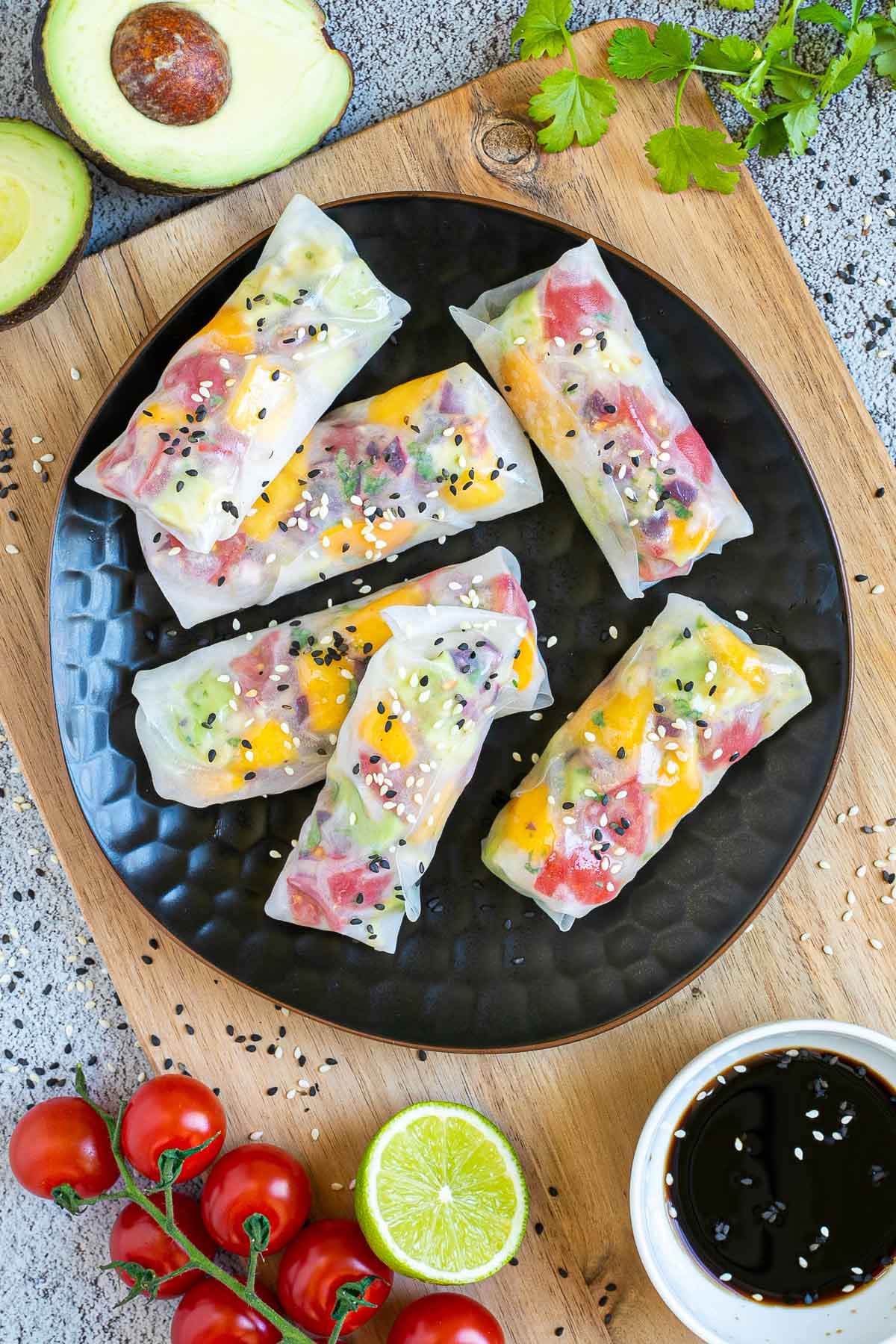 6 fresh rice paper wraps filled with chopped tomato, mango, and avocado placed on a black plate. Sprinkled with white and black sesame seeds. Leftover fillings are next to them. with a black dipping sauce