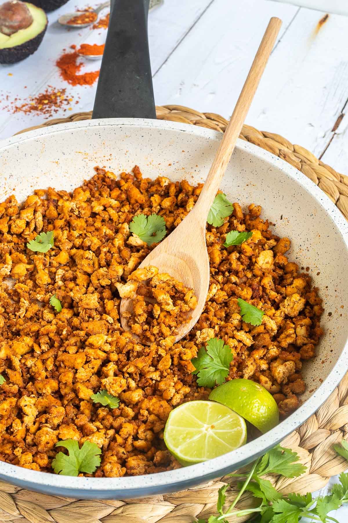 A white frying pan with brown crispy tofu crumbles sprinkled with freshly chopped cilantro. Lime slices and a wooden spatula are added. Avocado and more spices are next to the pan.