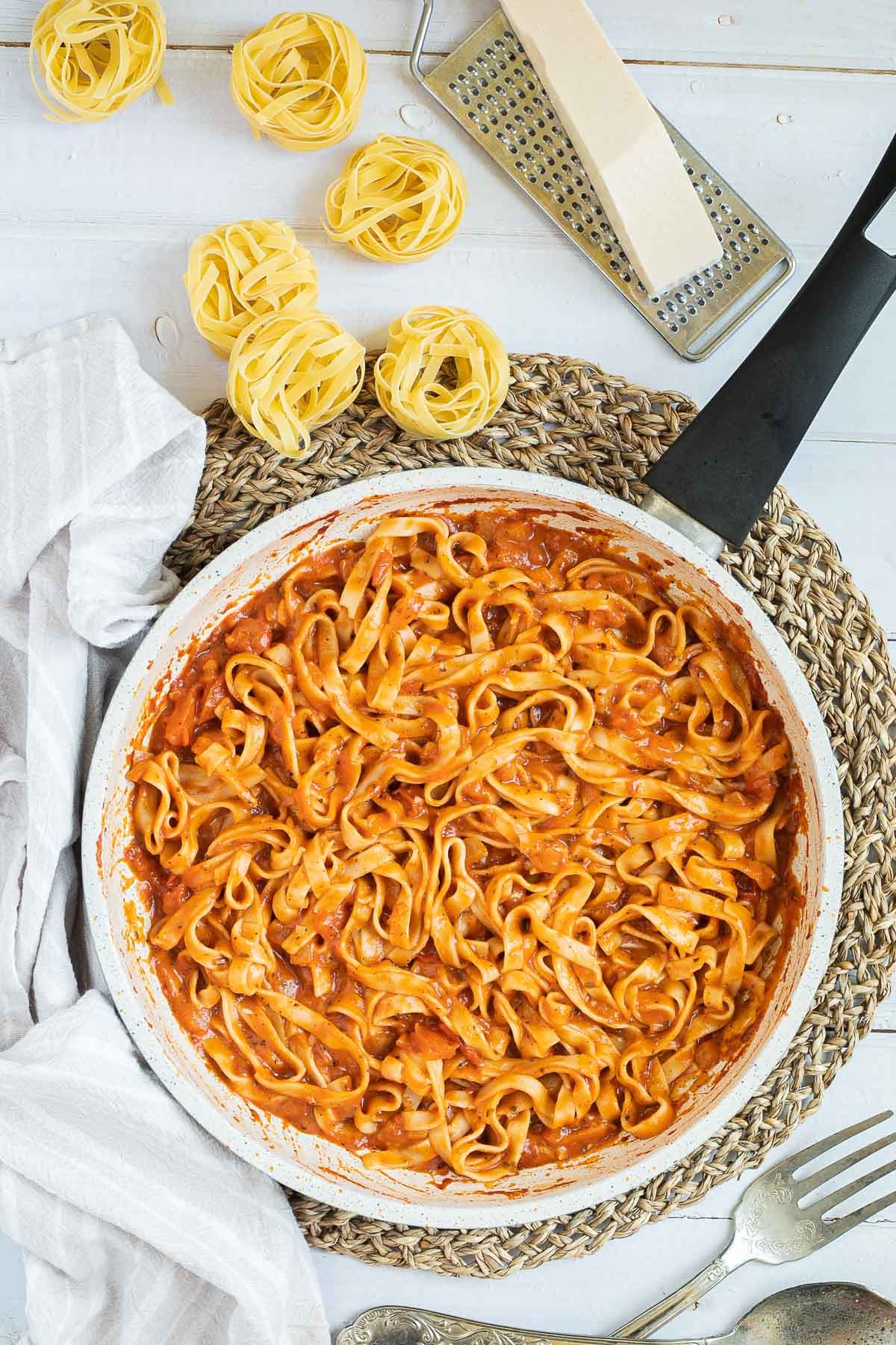 Tagliatelle is mixed in and covered with a creamy light red tomato-based sauce in a white frying pan. Dry tagliatelle pasta and a cheese grater with parmesan cheese is next to it.