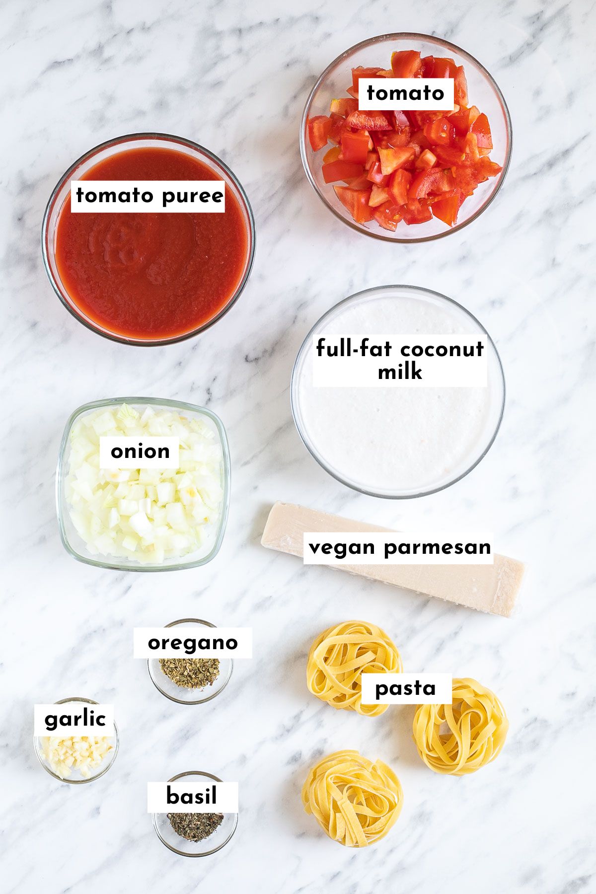 The ingredients of coconut milk pasta sauce is in small glass bowls like tomato sauce, chopped tomato, chopped onion, coconut cream, chopped garlic, dried green herbs, a piece of parmesan cheese and some dry tagiliatelle pasta.