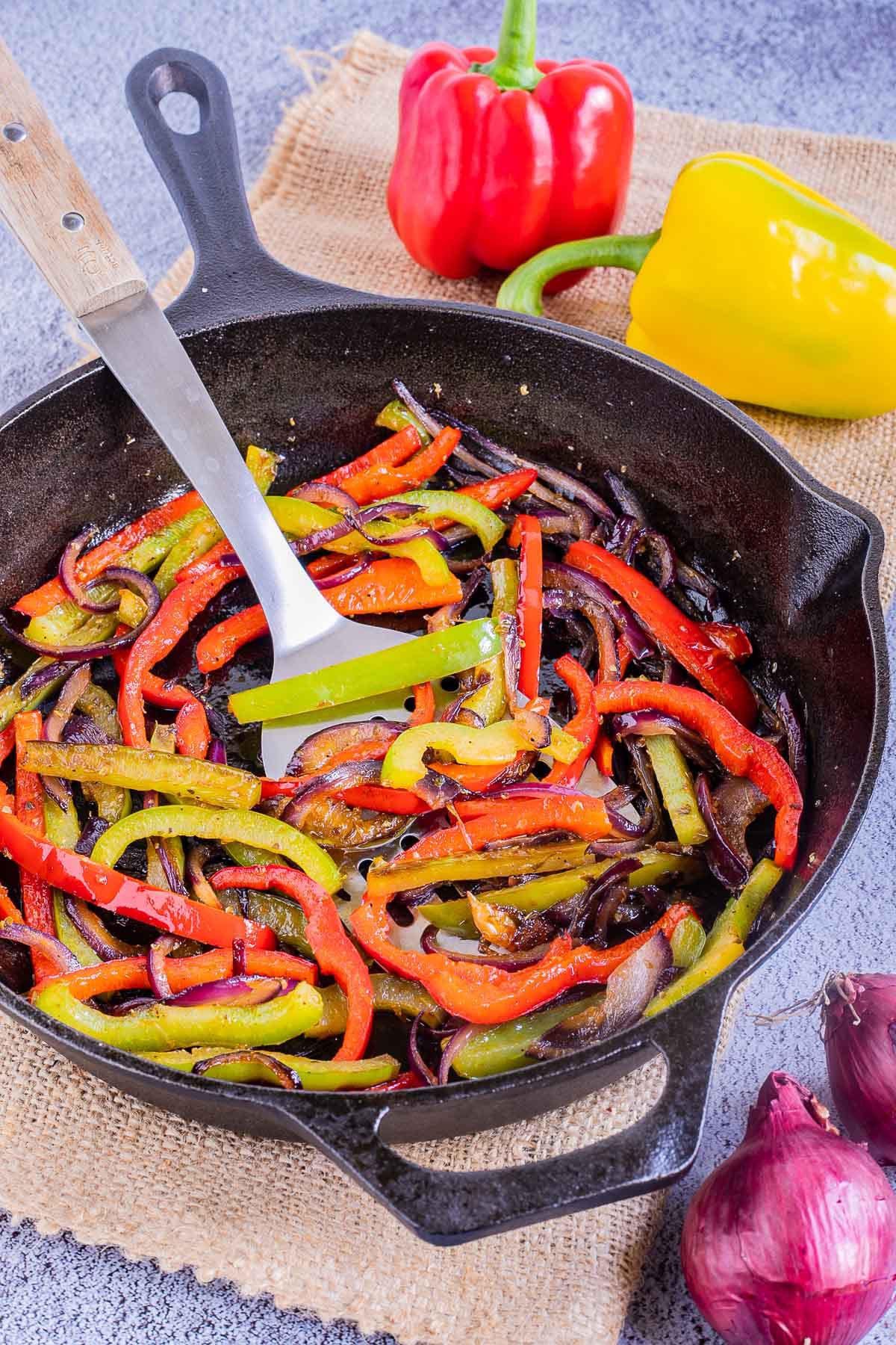 Black cast iron skillet with slightly brown red and green bell pepper strips and red onion slices. A slotted turner is place in the middle.