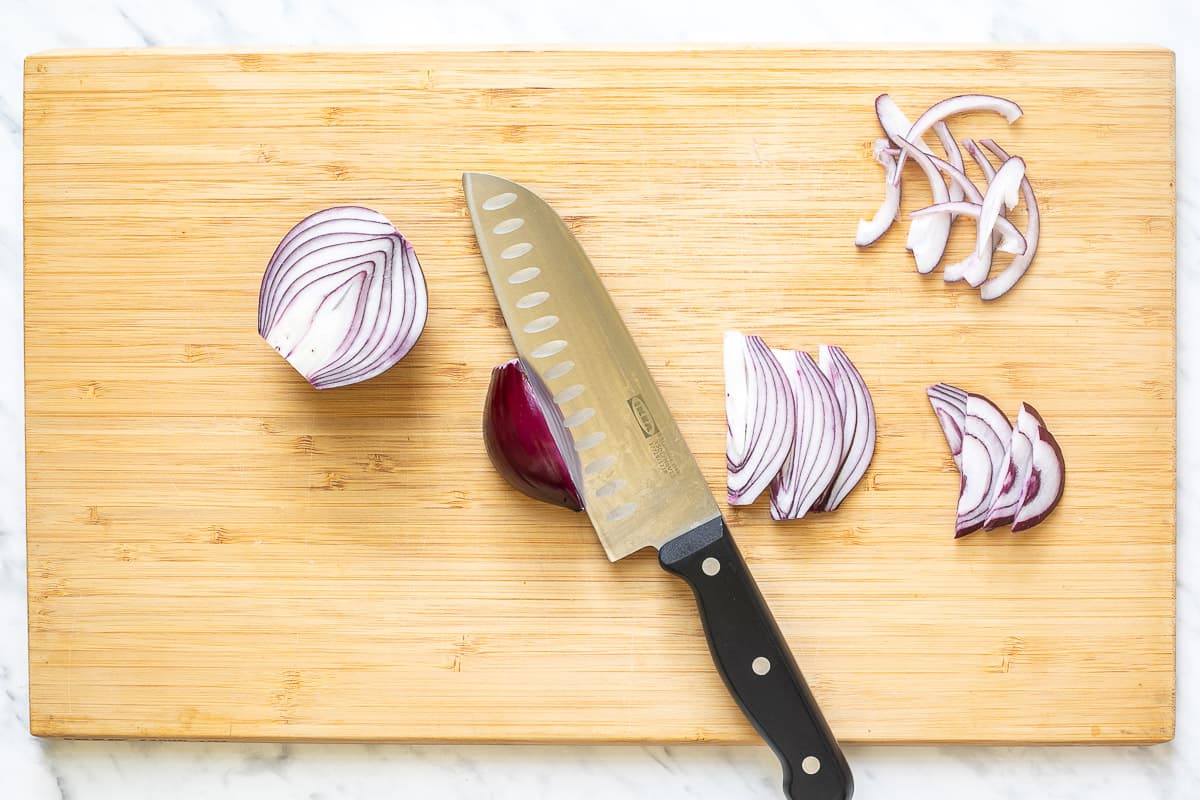 A wooden cutting board with a knife and red onion slices and halves.