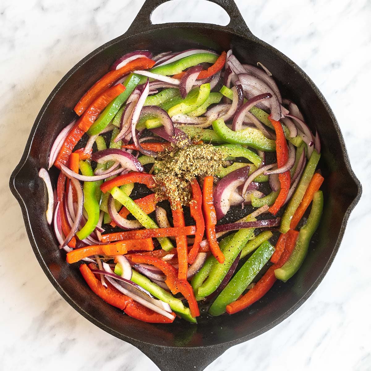 Black cast iron skillet with slightly brown red and green bell pepper strips and red onion slices, oregano on top.